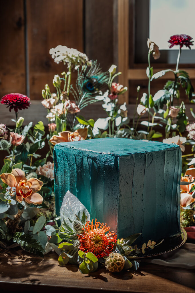 Square wedding cake with dark teal frosting. Colorful floral arrangement around the cake. 