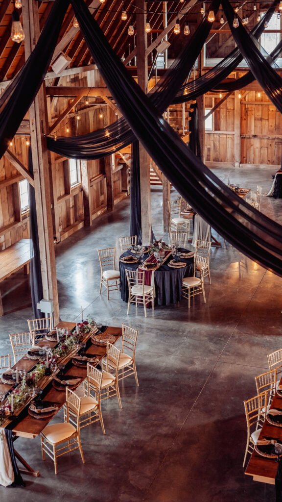 Barn wedding venue with string lights across the ceiling. Black drapes hang from ceiling. Room has a moody aesthetic.