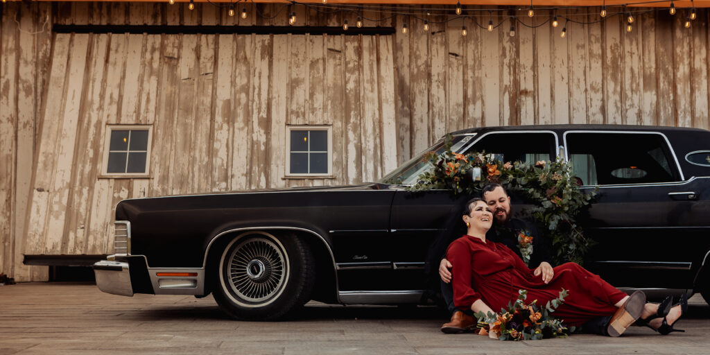 Couple sitting in front of black car with a floral installation piece on the door of the car. 